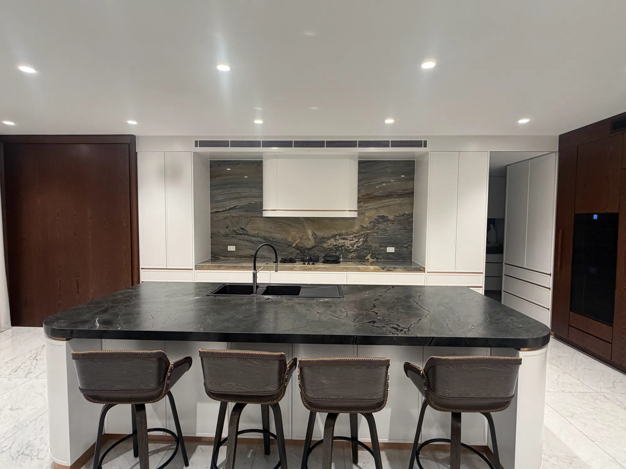 White kitchen with a dark stone island and four stools, a veined stone splashback behind a curved white rangehood, and dark timber tall cabinets
