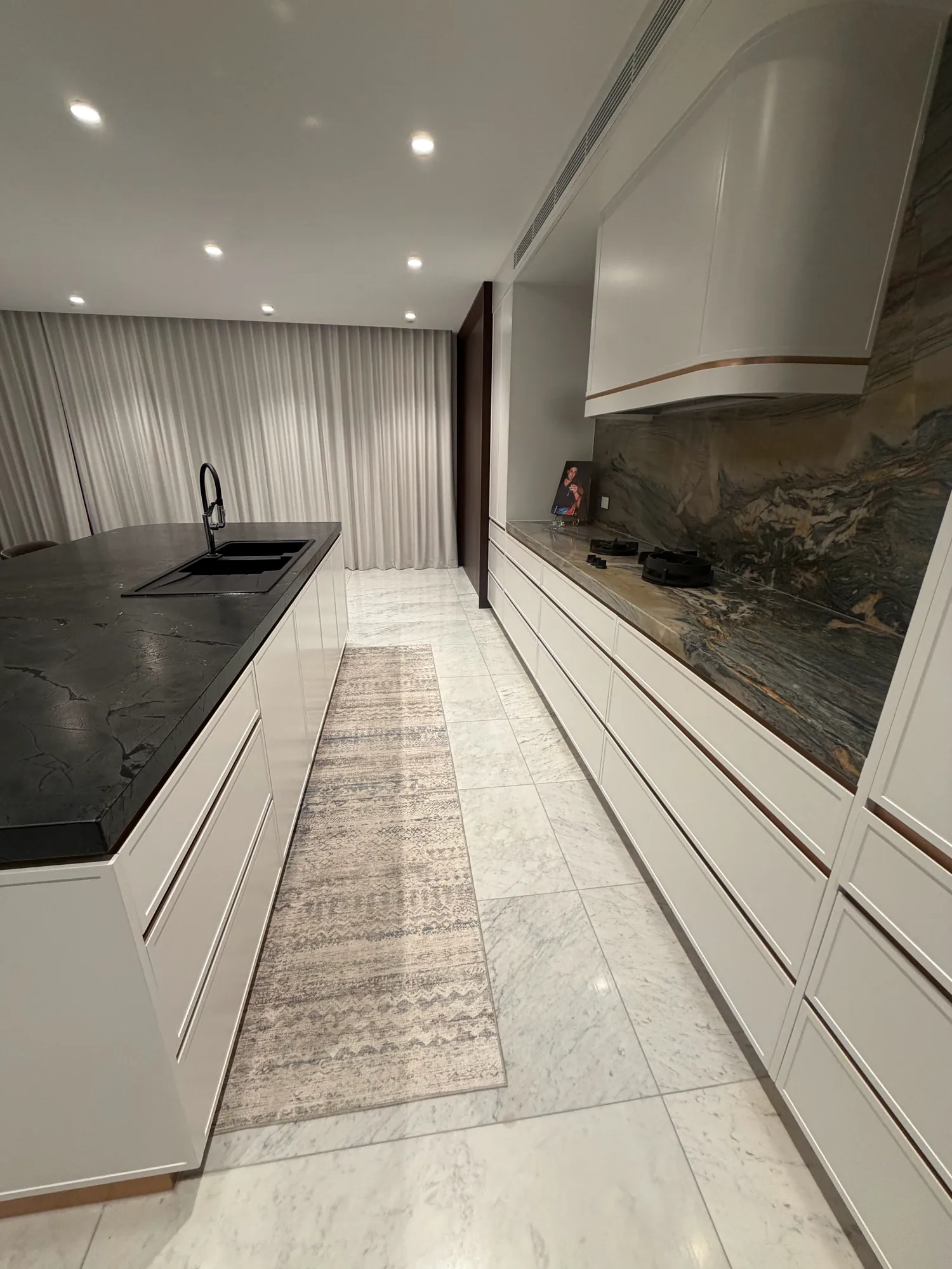 View down a kitchen between the dark stone island and a run of white drawers with a stone benchtop and splashback