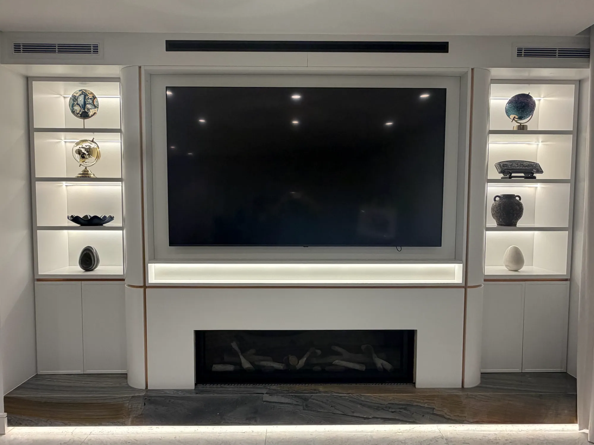 White built-in entertainment unit with a recessed television, a fireplace below and lit display shelves either side